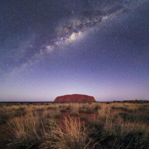 Uluru at night with the milky way shining above it in the deep purple sky. 