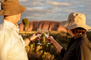 Two people toasting over a class of champagne as they look out at Uluru in the distance